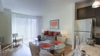 This single-floor space combines a living area and dining space in an open-concept layout that promotes a sense of airiness and flow. Large floor-to-ceiling windows dressed with vertical blinds allow abundant natural light to fill the room, creating a bright and welcoming atmosphere throughout.

The living area is anchored by a contemporary gray sofa accented with bold red throw pillows, complemented by a sleek black coffee table and a neutral armchair. A framed piece of wall art adds a pop of color, reinforcing the modern aesthetic. Ambient lighting is provided by stylish table lamps positioned on either side of the seating arrangement.

Adjacent to the living area, the dining space features a round glass-top table paired with upholstered chairs, adding an elegant yet functional touch. Red placemats echo the color palette established in the living zone, creating visual cohesion across the open floor plan.

Warm hardwood-style flooring runs throughout, grounding the space and contrasting pleasantly with the crisp white walls and ceiling. A partial view of stainless steel kitchen appliances hints at a connected kitchen area, completing this well-appointed, thoughtfully designed space.