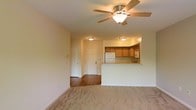 This single-story space offers an open-concept layout that connects the living area and kitchen, creating a functional and cohesive floor plan. The living area is finished with neutral-toned carpet, providing a comfortable underfoot feel, while a ceiling fan with a built-in light fixture serves as a central overhead feature, adding both practicality and a modest design element to the room.

The kitchen is positioned at the back of the space and is defined by a raised breakfast bar counter, which acts as a natural divider between the cooking and living zones while maintaining the open feel. Warm medium-toned wooden cabinetry lines the kitchen walls, offering ample storage. A white refrigerator and what appears to be a built-in microwave are among the included appliances. The kitchen area transitions to hardwood-style flooring, contrasting with the carpeted living space.

Two doors are visible toward the back of the unit — likely leading to an entryway and additional rooms. Flush-mount light fixtures illuminate the kitchen and hallway areas. The overall ambiance is clean, neutral, and move-in ready, with a straightforward and practical design throughout.