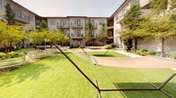 The Fire Pit and Hammock Courtyard is a thoughtfully designed outdoor amenity space nestled within a multi-story residential community. Enclosed on multiple sides by building facades with private balconies and ground-level patios, the courtyard offers a sense of intimacy while remaining open to the sky above. The layout is anchored by a circular fire pit surrounded by vibrant pink metal chairs, creating an inviting focal point for social gatherings. Lush, mature trees are planted within large galvanized steel circular planters, adding an industrial-meets-natural aesthetic throughout the space. A decomposed granite pathway winds through the courtyard, flanked by well-maintained native and ornamental plantings including agave, ferns, and ground cover shrubs. A generous lawn area provides space for relaxation, with freestanding hammocks offering a leisurely retreat. Cornhole boards are stationed along the pathway, adding a recreational element to the outdoor experience. Ground-level units feature private patios enclosed by sleek metal railings, seamlessly integrating residential living with the shared courtyard environment. Overall, the space balances active recreation with serene green space in a well-landscaped, community-centered design.