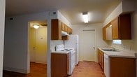 This single-floor residential unit offers a practical and well-organized layout designed for comfortable everyday living. The kitchen serves as a central feature of the space, outfitted with warm honey-toned wood cabinetry that extends to the upper walls for ample storage. White appliances — including an electric coil range, refrigerator, and dishwasher — provide a clean, classic look that complements the neutral wall tones. A range hood is mounted beneath the upper cabinets, and overhead fluorescent lighting ensures the workspace is well lit. The kitchen flows seamlessly into the adjoining areas via an open passageway, offering an interconnected feel throughout the unit. Rich hardwood flooring runs through the kitchen and into the adjacent hallway, adding warmth and visual continuity. The hallway beyond the kitchen is softly lit with a ceiling-mounted light fixture and features a door leading to additional rooms within the unit. Ventilation is addressed through ceiling-mounted air vents visible throughout the space. The overall ambiance is functional and homey, with a straightforward design that prioritizes usability and a welcoming, lived-in character.