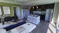 This single-floor residence showcases a thoughtfully designed open-concept layout that seamlessly integrates the living, dining, and kitchen areas into one cohesive, flowing space. The kitchen is a standout feature, outfitted with sleek dark cabinetry, stainless steel appliances, a mosaic tile backsplash, and a central island with a built-in sink and breakfast bar seating accommodating multiple stools — ideal for casual dining and entertaining. Pendant lighting and a track lighting system provide layered illumination throughout the space, enhancing both functionality and ambiance. The living area is anchored by a clean-lined white coffee table and a deep blue sofa, complemented by a white cube shelving unit that adds storage and visual interest. Rich, dark hardwood flooring runs throughout, grounding the space and providing a striking contrast against the lighter walls accented with warm green tones. A sitting nook with a leather armchair adds a cozy, private corner to the otherwise open layout. Interior doors lead to additional rooms, offering a sense of depth and functionality. The overall design balances modern aesthetics with practical livability, making it an inviting and well-appointed space.