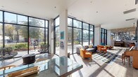 Located in Houston, TX (77002), this striking single-floor space is defined by its modern architectural design and an exceptional sense of openness. Floor-to-ceiling glass curtain walls with black steel framing wrap around the perimeter, flooding the interior with natural light and offering panoramic views of the surrounding streetscape and mature trees outside.

The main gathering area features wide-plank wood flooring, exposed concrete structural columns, and soaring ceiling heights that create an airy, gallery-like atmosphere. Eclectic seating arrangements — including navy geometric-patterned area rugs, orange accent chairs, a gray sectional sofa, and vibrant teal lounge chairs — provide multiple comfortable zones throughout the open-plan layout.

The space doubles as an art venue, with an extensive rotating display of colorful paintings, mixed-media works, and framed pieces adorning the white walls. A classic upright piano anchors one corner, reinforcing the creative and cultural character of the space.

A dedicated kitchenette area features light wood cabinetry, granite countertops, a penny-tile backsplash, and a retro-styled SMEG refrigerator, providing functional refreshment facilities. A secondary seating and dining area with natural wood furnishings offers additional gathering space.

Glass partitions and black-framed doors connect interior zones while maintaining visual transparency throughout, making this a dynamic, light-filled space ideal for community, cultural, or creative use.