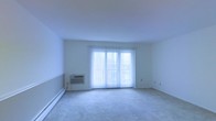 This single-floor space offers a clean, minimalist layout designed for comfortable everyday living. The main living area is generously sized, featuring wall-to-wall carpeting that adds warmth underfoot and a neutral color palette of crisp whites throughout the walls and ceiling, creating a bright and airy atmosphere. Large sliding glass doors serve as a focal point of the room, fitted with vertical blinds that allow for adjustable natural light and privacy. The doors open to an outdoor view, bringing in abundant daylight that fills the entire space. A wall-mounted air conditioning unit is conveniently positioned near the sliding doors, ensuring climate control throughout the warmer months. Supplementing the heating system, a baseboard heater runs along the perimeter of the room, providing efficient warmth during cooler seasons. The overall ambiance of the space is light, open, and functional — ideal for those seeking a straightforward, move-in-ready layout. The simple, uncluttered design allows for flexible furniture arrangement and personalization to suit a variety of lifestyles and needs.