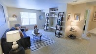 This single-floor space offers a comfortable and thoughtfully designed living environment. The living area serves as the centerpiece of the home, featuring plush wall-to-wall carpeting that adds warmth underfoot. A large window floods the room with natural light, creating a bright and inviting atmosphere throughout the day. Built-in wall shelving provides both functional storage and an opportunity for tasteful display, complemented by freestanding ladder shelves that add a stylish, contemporary touch. The neutral gray walls offer a versatile backdrop that pairs well with a range of furnishing styles. A dedicated wall niche with a built-in desk or ledge adds a practical element to the living space. The open layout allows for flexible furniture arrangement, as demonstrated by the centrally placed coffee table and coordinated seating area anchored by a striped area rug. Soft, ambient lighting from a table lamp near the entryway enhances the warm, welcoming feel of the space during evening hours. Overall, the design balances functionality with a clean, modern aesthetic, making it a practical and comfortable place to live.