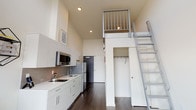 Located on the 7th floor at 5001 Brooklyn Avenue NE in Seattle's University District, this loft-style unit offers a striking vertical design with soaring ceilings that create an open, airy atmosphere. A metal ladder leads to an elevated sleeping loft enclosed by a sleek metal railing, maximizing the use of vertical space while adding architectural character to the interior.

The main living area features rich, dark hardwood flooring throughout, complementing the crisp white walls and modern aesthetic. The well-appointed kitchen is fitted with white flat-panel cabinetry, a tile backsplash, built-in cooktop, sink, stainless steel microwave, and a full-size refrigerator — providing all essential cooking amenities in a clean, contemporary layout.

A large picture window in the living area floods the space with natural light while framing panoramic views of the surrounding Seattle cityscape, offering an engaging urban backdrop. A built-in closet with rod and shelving provides convenient storage just off the main living area.

The bathroom is outfitted with a full soaking tub and shower combination featuring built-in corner shelving, finished in clean white surrounds with chrome fixtures. The sleeping loft above benefits from the same large window views, creating a bright and functional retreat elevated above the living space below.