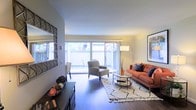 This thoughtfully designed single-floor space offers a welcoming and comfortable atmosphere, highlighted by an open-concept living area that maximizes both functionality and style. The living room serves as the heart of the space, featuring rich hardwood flooring that runs throughout and lends a warm, grounded feel to the interior. A large set of sliding glass doors spans nearly the full width of the room, flooding the space with abundant natural light and offering direct access to a private outdoor area enclosed by a wood-panel fence, providing a sense of seclusion and greenery. The interior is complemented by smooth, neutral-toned walls and a clean white ceiling that together create a bright and airy ambiance. A decorative geometric mirror on the wall adds a stylish architectural accent, while the overall layout provides ample room for both lounging and entertaining. The transition between indoor and outdoor living is seamless, making the space feel more expansive than its footprint suggests. The combination of natural light, quality finishes, and thoughtful design elements creates an inviting environment that is both practical and aesthetically refined.