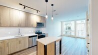 Located at 2311 Dulles Station Blvd in Herndon, Virginia, this modern single-floor residence offers a thoughtfully designed open-concept layout with contemporary finishes throughout. The kitchen is a standout feature, showcasing light wood cabinetry, white quartz countertops, a subway tile backsplash, and a full suite of stainless steel appliances including a refrigerator, electric range, microwave, and dishwasher. A freestanding island with a white top and dark metal legs anchors the space, while stylish pendant lighting and a track lighting system illuminate the area with a clean, modern aesthetic. Light wood-toned luxury vinyl plank flooring extends seamlessly from the kitchen into the open living area, which benefits from large floor-to-ceiling windows that flood the space with natural light and offer views of the surrounding community. The bedroom is carpeted for comfort and features multiple windows with blinds, providing ample natural light. A generously sized walk-in closet is outfitted with multi-level wire shelving and hanging rods, offering excellent storage capacity. The bathroom is elegantly appointed with warm wood-toned cabinetry, a white quartz vanity countertop, a large illuminated mirror, and a glass-enclosed tile shower, creating a refined and functional space.