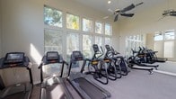 This single-story fitness center is designed to support a comprehensive workout experience within a bright, open environment. The space is defined by its soaring ceilings and expansive floor-to-ceiling windows that line multiple walls, flooding the interior with natural light and offering views of surrounding palm trees and greenery. Multiple ceiling fans are mounted throughout, ensuring consistent airflow across the entire floor.

The cardio zone is well-stocked with Matrix-brand equipment, including treadmills, elliptical trainers, stair climbers, and upright stationary bikes, all positioned to take advantage of the outdoor views. The flooring throughout is a durable, speckled carpet-and-rubber composite suited for high-traffic fitness use.

A dedicated functional training area occupies one section of the room, outfitted with a multi-station cable and rack system, kettlebells, medicine balls, and TRX suspension training equipment. Full-length mirrors line portions of the walls, enhancing the sense of space and supporting form-focused training.

Near the entrance, practical amenities include a water cooler station, a bulletin board with posted information, and a fire extinguisher. Blinds on the windows allow light control as needed. The overall ambiance is clean, airy, and well-maintained, creating an inviting environment for a wide range of fitness activities.