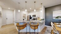 This thoughtfully designed single-floor residence offers 2 bedrooms and 2 bathrooms across a comfortable and functional layout. The kitchen is a standout feature, showcasing crisp white shaker cabinetry with matte black hardware, granite countertops, and a full suite of stainless steel appliances including a refrigerator, range, and microwave. A peninsula with seating for two is illuminated by a row of four pendant lights with brass accents, making it an ideal casual dining spot. The adjacent dining area provides a more formal setting, framed by a charcoal accent wall that adds depth and character to the space. The living room offers natural light through window treatments and warm wood-style flooring that flows throughout the home. The primary bedroom is generously sized, featuring a ceiling fan, large windows with outdoor views, and ample room for furnishings. The second bedroom is equally comfortable. Both bathrooms are finished with white vanity cabinetry and granite countertops, consistent with the home's overall design palette. A dedicated laundry room houses a full-size washer and dryer with built-in shelving for added storage. The cohesive aesthetic throughout—light tones, wood-style flooring, and quality finishes—creates a bright and inviting living environment.