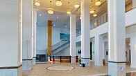 Located along the waterfront in Fort Myers, Florida, this impressive commercial lobby at 2500 Edwards Dr offers a grand and polished environment designed to make a lasting first impression. The space is defined by its dramatically soaring ceilings, supported by a series of tall, marble-clad columns with illuminated vertical light strips that draw the eye upward and lend a sophisticated, modern ambiance throughout.

The main atrium features large-format beige tile flooring with a striking circular marble inlay as a central focal point. Floor-to-ceiling glass walls and expansive windows flood the interior with abundant natural light while offering sweeping views of the surrounding waterfront and lush outdoor greenery. A seating lounge near the glazed façade provides a welcoming area furnished with elegant white sofas and a glass coffee table.

A prominent staircase with granite treads and sleek cable railings connects to an upper mezzanine level, which overlooks the atrium below. The lobby also features a stylish reception desk with warm wood paneling and accent lighting, a vibrant living green wall, and an integrated café — Casanova — complete with a display case and espresso bar. Multiple hallways extend from the main atrium, guiding visitors through the space with ease. The overall atmosphere is refined, bright, and professional.