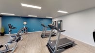 This fitness center offers a clean, modern environment designed to support an active lifestyle. The space is laid out on a single floor, featuring light wood-style flooring throughout that adds warmth to the otherwise crisp, neutral interior. Bright recessed panel lighting illuminates the room evenly, complemented by natural light streaming in through multiple windows that offer views of the surrounding neighborhood.

The cardio equipment area is well-stocked, housing multiple treadmills, upright and recumbent stationary bikes, and elliptical machines, all arranged to allow comfortable use and circulation. An exposed industrial-style duct runs along the ceiling near the window wall, adding a subtle modern-industrial touch to the space. A wall-mounted flat-screen television is positioned for easy viewing during workouts.

A bold teal accent wall on one side of the room provides a vibrant contrast to the otherwise neutral white and gray palette, adding visual energy to the environment. A wall clock and a small high-top table with seating offer practical convenience for users. Glass-paneled interior doors and windows connect the fitness area to adjacent spaces, maintaining an open and airy feel. A stainless steel waste bin and tidy layout reflect the well-maintained nature of the facility.