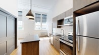 Located at 2929 Main St in Buffalo, NY, this thoughtfully designed single-floor unit blends industrial character with modern finishes throughout. The open-concept kitchen serves as the centerpiece of the space, featuring sleek taupe cabinetry that extends to the ceiling, a classic white subway tile backsplash, dark countertops, and a full suite of stainless steel appliances including a range, built-in microwave, dishwasher, and refrigerator. A kitchen island with a dark countertop provides additional workspace and casual seating. Dramatic black pendant lights hang from the soaring ceilings, reinforcing the space's loft-like aesthetic, while a track lighting system adds functional illumination. Light hardwood flooring flows throughout the main living areas, complemented by exposed structural columns that add to the industrial character. The bedroom areas feature carpeted flooring and oversized double-hung windows framed by painted brick walls, flooding the rooms with natural light and offering views of the surrounding neighborhood. The bathroom is appointed with warm wood cabinetry, a dark countertop vanity, a large mirror, and a full tub-shower combination, all finished with brushed nickel hardware and light hardwood-style flooring.