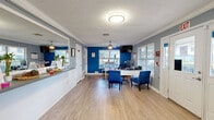 Located at 3780 University Club Blvd in Jacksonville, FL, this single-floor commercial space functions as a welcoming reception and waiting area. The layout includes a distinct entrance and a combined living/reception room, making efficient use of its compact footprint.

The interior is defined by a striking two-tone color palette — bold cobalt blue accent walls contrast with soft gray surroundings — creating a vibrant yet professional atmosphere. Light wood-look flooring runs throughout, complementing the white crown molding and wainscoting details that add a polished, finished look. A textured ceiling with flush-mount lighting and ceiling fans ensures consistent airflow and illumination across the space.

The reception area features a built-in counter with a large pass-through window, ideal for staff-to-visitor interaction. Display items and greenery on the counter add a welcoming touch. Adjacent to the counter, a dedicated workstation with a desk, monitor, and blue upholstered chairs provides a functional office area. Multiple windows with horizontal blinds allow natural light to filter in while maintaining privacy.

The waiting area is furnished with a mid-century modern sofa, a patterned area rug in blue and gold tones, and a gold-accented console table with a coffee station. A wall-mounted television and a bird cage add unique character to the space. The entrance features a white paneled door with sidelight windows, offering views of the surrounding outdoor greenery.