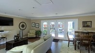 This single-story space is designed with a warm, inviting aesthetic that balances comfort and functionality. The main living area is anchored by rich hardwood flooring that contrasts elegantly with the soft, neutral-toned walls finished in warm beige. Crown molding frames the room with a classic architectural detail, while recessed lighting in the white ceiling provides clean, even illumination throughout. A decorative tray ceiling detail adds a subtle architectural accent overhead.

The room's most prominent feature is a set of white-framed French doors that span much of the exterior wall, flooding the space with abundant natural light and offering direct views of lush greenery and outdoor surroundings. These doors create a seamless visual connection between the interior and the outdoors.

The living area is generously proportioned, accommodating a substantial seating arrangement including a sofa and armchair in neutral upholstery, complemented by a patterned area rug. Wall art and a large decorative clock contribute character to the space. A dining area is partially visible to one side, suggesting a practical open-plan layout ideal for everyday living and entertaining.