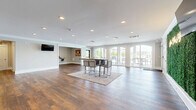Located at 200 W Edge Way in Lynchburg, Virginia, this single-floor community clubhouse offers a well-appointed and versatile layout designed for both relaxation and activity. Rich hardwood-style flooring flows seamlessly throughout, creating a cohesive and polished aesthetic across all areas.

The two living rooms serve distinct purposes while sharing a bright, open character. The primary gathering space features a central high-top table with bar seating, recessed lighting, a wall-mounted flat-screen TV, and a lush green living wall accented by a "Good Vibes Only" neon sign — adding a lively, welcoming energy. A row of large arched floor-to-ceiling windows and glass doors lines one wall, flooding the room with natural light and offering direct access to an outdoor patio furnished with seating. A community bulletin board in the secondary living area serves as an informational hub for residents.

The dedicated exercise room is enclosed by full-length glass panel walls and doors, providing visibility into the well-equipped fitness space while maintaining a sense of separation. Mirrors line the interior walls, and the room is outfitted with cardio and strength-training equipment. Crown molding and recessed lighting throughout elevate the overall finish of the space, while the connecting hallway ties all areas together efficiently.