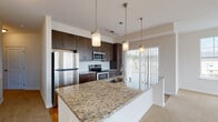 This single-floor residence offers a thoughtfully designed open-concept layout that seamlessly connects the living area and kitchen. The living space is finished with plush wall-to-wall carpeting and benefits from abundant natural light through a large multi-pane window, with views of the surrounding neighborhood. A ceiling fan adds both function and comfort to the area, while sliding glass doors provide access to an outdoor balcony.

The kitchen is a standout feature, showcasing rich dark-wood cabinetry that extends to the ceiling, a classic white subway tile backsplash, and a generous granite-topped peninsula with an undermount sink — ideal for both meal prep and casual dining. Stainless steel appliances, including a refrigerator, electric range, and built-in microwave, complete the space. Warm hardwood-style flooring anchors the kitchen area.

The bedrooms are carpeted and well-lit, with one featuring a large window overlooking a green outdoor setting. A dedicated walk-in closet offers extensive wire shelving for ample storage. The bathroom is finished with hardwood-style flooring, a granite vanity with dark cabinetry, and a glass-enclosed walk-in shower with white tile surround. A dedicated laundry room accommodates a full-size washer and dryer with additional wire shelving above.