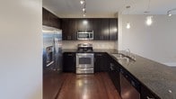 This single-floor residence showcases a thoughtfully designed open layout, anchored by rich dark hardwood flooring that flows seamlessly throughout the main living areas. The open-concept living space is generously proportioned, featuring a ceiling fan, track lighting, and direct access to a private balcony through glass doors that invite natural light into the room.

The kitchen is a standout feature, outfitted with dark espresso cabinetry, granite countertops with a subtle speckled finish, and a full suite of stainless steel appliances including a French door refrigerator with ice maker, a range with oven, built-in microwave, and dishwasher. A tile backsplash adds texture, while pendant lights and track lighting illuminate the workspace. A kitchen island with a dark granite top provides additional prep space and separates the kitchen from the living area.

A hallway leads to a walk-in closet equipped with multi-tiered wire shelving and hanging rods, offering ample storage. The bathroom features large-format tile flooring, a tiled tub-and-shower combination, a dark vanity with a white countertop, and open shelving for added functionality. The overall aesthetic is clean, contemporary, and well-maintained throughout.