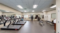 Located at 9000 Stebbing Way in Laurel, Maryland, this fitness center offers a comprehensive, well-equipped workout facility designed to support a variety of exercise routines. The space is housed on a single floor and features a spacious, open layout with a drop-panel ceiling, recessed fluorescent lighting, and ceiling fans that contribute to a comfortable training environment. Durable rubber-textured flooring covers the entire workout area, providing safety and support for all types of exercise. Large mirrors line multiple walls, enhancing the sense of space and allowing users to monitor their form. Natural light filters in through several windows, keeping the atmosphere bright and welcoming. The cardio section is well-stocked with multiple treadmills, elliptical machines, and a stationary bike. The strength training area includes a multi-function cable machine, a dumbbell rack with a full range of free weights, adjustable weight benches, and a rowing machine. A wall-mounted flat-screen television is positioned for easy viewing during workouts. Additional amenities include rolled exercise mats, stability balls, and conveniently placed storage shelving. The facility's clean, neutral-toned walls and organized layout create a focused and functional environment suited for a broad range of fitness goals.