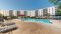 Located at 400 Foulk Rd in Wilmington, Delaware, this outdoor amenity space serves as a central gathering area for the surrounding multi-story residential community. The centerpiece is a generously sized, freeform swimming pool with crystal-clear blue water, designed with a curved silhouette that adds a resort-like character to the setting. The pool deck is finished in a smooth concrete surface and offers ample space for lounging, furnished with a well-appointed selection of chaise lounge chairs, wicker seating, and shade umbrellas — including a bold red patio umbrella that adds a pop of color to the scene. A smaller secondary pool or spa area is also present near the lounge seating, providing an additional relaxation option. The entire pool area is enclosed by an iron perimeter fence, ensuring privacy and security while maintaining open sightlines to the lush mature trees and manicured landscaping that border the space. Ornamental grasses and seasonal plantings add natural texture throughout. A small utility or pool house structure is situated at the edge of the deck, complementing the functional layout. The backdrop of the multi-wing residential building, featuring balconies on every floor, frames the outdoor space and reinforces the community-centered design of this well-maintained property.
