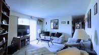 This single-floor residence centers around a spacious living room that serves as the heart of the home. Large vertical-blind-covered windows allow abundant natural light to fill the room, creating a bright and airy atmosphere. The living room is furnished with a white upholstered sofa and matching armchairs arranged around a sleek glass-and-metal coffee table, all set atop a light-colored area rug. Decorative wooden sculptures and framed artwork on the walls add cultural character to the space. A flat-screen television is mounted on an entertainment unit along one wall, accompanied by a media storage shelf. A window-mounted air conditioning unit and a standing fan provide climate control. Lush indoor plants in the corner introduce a natural element. A hallway visible to the right suggests additional rooms beyond the living area, with the corridor displaying further framed artwork along its walls. The overall ambiance is comfortable and lived-in, with a mix of functional furnishings and personal decorative touches that give the space a warm, personalized character.
