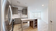 This single-floor residence showcases a thoughtfully designed open-concept layout with a clean, contemporary aesthetic. The kitchen serves as a focal point, featuring taupe shaker-style cabinetry, a marble-look tile backsplash, and quartz countertops. A central island with built-in shelving provides additional storage and casual seating, while stainless steel appliances—including a French door refrigerator, microwave, and dishwasher—complete the well-appointed space. Under-cabinet lighting enhances both function and ambiance.

Light hardwood-style flooring extends throughout the main living areas, reflecting natural light and contributing to the unit's bright, airy feel. The open living area flows seamlessly from the kitchen, offering a flexible layout with recessed lighting overhead. A door with large glass panels provides access to a private outdoor balcony, allowing natural light to filter into the space.

The bedroom features plush carpeting, a ceiling fan, and French doors leading to an outdoor balcony, creating a comfortable and well-ventilated retreat. The bathroom is finished with a backlit vanity mirror, quartz countertop, taupe cabinetry with gold hardware, wood-look tile flooring, and a glass-enclosed walk-in shower with tiled walls, delivering a polished and functional design.