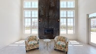 Located at 12822 White Willow Dr in Plainfield, Illinois, this single-floor community clubhouse is designed to impress with a bright, open layout and thoughtfully curated architectural details. The entrance welcomes visitors through an elegant arched colonnade, where crisp white walls, light-toned tile flooring, and soaring ceilings create an immediately airy and inviting atmosphere. A central seating area features a mid-century modern tufted blue loveseat and colorful area rug, setting a warm and contemporary tone throughout.

The standout feature of the main lounge is a dramatic, floor-to-ceiling fireplace clad in a richly textured oxidized metal surround, which rises to meet the full height of the room. Flanking the fireplace on either side, expansive multi-pane windows flood the interior with generous natural light and frame views of the outdoor pool area and surrounding community. Two floral accent chairs and a marble-topped side table complete the intimate fireside seating arrangement.

Glass double doors lead directly to the outdoor pool terrace, seamlessly connecting the interior to the exterior amenity space. A front desk or reception area is also visible within the layout, suggesting this space serves as a central hub for community activity. The overall ambiance is clean, modern, and welcoming — balancing stylish design elements with practical, community-focused functionality.