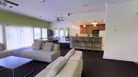 This single-floor space is designed as an open-plan gathering area that seamlessly integrates a lounge, dining zone, and full kitchen. The lounge area anchors the space with two generously sized neutral sofas accented with patterned throw pillows, arranged to encourage comfortable conversation. Dark carpeting grounds the living area and contrasts with the bright white walls adorned with distinctive green arched alcoves, lending a playful architectural character to the room. A ceiling fan overhead aids air circulation throughout the space. Large windows along one wall flood the dining area with abundant natural light, illuminating a cluster of dining tables and high-back chairs suited for group gatherings. The kitchen, positioned toward the rear, features warm wood cabinetry, a built-in microwave, and a white refrigerator. A breakfast bar with bar-height seating provides additional casual dining options. Pendant lights hang above the bar area, adding a warm, focused glow. The overall ambiance is bright, functional, and communal, making this space well-suited for social gatherings, meetings, or shared meals.