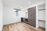 This single-floor unit showcases a clean, contemporary design throughout, characterized by crisp white walls, recessed lighting, and light-toned hardwood-style flooring that runs consistently across the living spaces. The open layout connects the main living area to a well-appointed kitchenette, which features flat-front cabinetry in a warm wood-grain finish, a white countertop with an undermount sink, matte black fixtures, an induction cooktop, a compact refrigerator, and a microwave tucked neatly into a dedicated cabinet opening. A globe pendant light hangs above the kitchenette, adding a modern accent, while a window above the sink allows natural light to filter in and provides an exterior view. The bedroom area is generously proportioned, offering clean walls and a dedicated walk-in closet alcove that provides practical storage. The bathroom is straightforward and functional, featuring an all-white walk-in shower stall with a curved base, bronze-finished fixtures, and a towel bar. The overall ambiance of the unit is bright, minimalist, and move-in ready, reflecting a thoughtfully designed modern aesthetic suited for efficient everyday living.