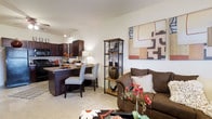 This single-floor residence showcases a thoughtfully designed open-concept layout that brings together the living and kitchen areas in a cohesive, welcoming environment. The living area is anchored by a plush sofa and accented with decorative touches, including a bold abstract canvas that adds a pop of color and artistic character to the neutral wall. A slim metal shelving unit serves as both a decorative and functional divider between the living and dining spaces, displaying curated décor pieces that enhance the room's warm aesthetic. Soft, neutral-toned carpeting runs throughout the living area, contributing to a cozy and comfortable atmosphere. The kitchen is outfitted with rich, dark wood cabinetry that creates a striking contrast against the light walls, paired with dark countertops that lend a sleek, modern feel. A peninsula with seating for two patterned accent chairs provides a casual dining option and serves as a natural gathering point. Pendant lighting overhead adds a stylish finishing touch to the kitchen. The overall ambiance of the space is warm and inviting, with a harmonious blend of dark wood tones, neutral backgrounds, and vibrant decorative accents that create a balanced and livable interior.