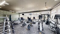 This fitness center in Baton Rouge, Louisiana offers a thoughtfully designed, single-floor workout facility that accommodates a comprehensive range of exercise options. The space features a clean, modern aesthetic defined by white walls accented with dark crown molding and trim, textured sage-green wall panels, and plush blue-gray carpeted flooring throughout.

Natural light floods the interior through an expansive bank of large, grid-pane windows lining multiple walls, complemented by a mix of recessed lighting, linear pendant fixtures, and ceiling fans overhead. A full-length mirror wall on one side amplifies the sense of openness and allows members to monitor their form during workouts.

The cardio zone is well-stocked with Matrix-brand treadmills, ellipticals, upright bikes, and rowing machines arranged in open rows facing the windows. The strength training area includes a cable machine, multiple weight benches, and a well-organized dumbbell rack with a wide range of weights. Medicine balls and additional functional training accessories round out the available equipment.

Wall-mounted televisions are positioned throughout the space for entertainment during workouts. Access is provided via elegant dark-framed glass doors, adding a polished touch to the overall design. The layout is efficient and spacious, allowing members to move freely between equipment stations without congestion.
