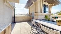 Located at 16101 Murphy Ave in Irvine, California, this rooftop terrace offers a sophisticated outdoor retreat designed for both relaxation and social gathering. The space is finished with large-format stone tiles throughout, lending a clean, polished aesthetic that complements the warm Southern California climate.

The terrace is thoughtfully divided into distinct functional zones. A dedicated lounge area features wicker seating with teal accent cushions arranged around low tables, providing a comfortable setting for casual gatherings. A covered pergola structure adds architectural interest while offering shaded seating beneath. A separate dining area with a bar-height table and metal chairs accommodates al fresco meals with open views of the surrounding skyline.

Tall freestanding pole lights are distributed across the terrace, ensuring the space remains well-lit and functional into the evening hours. Glass railings along the perimeter preserve unobstructed sightlines while maintaining safety, framing views of the lush palm trees and multi-story buildings of the Irvine area below.

Planters with manicured shrubs and tropical palms are strategically placed to add greenery and define the various seating zones. The open sky above and panoramic surroundings create a bright, airy ambiance, making this rooftop terrace a standout amenity that blends urban convenience with outdoor comfort.