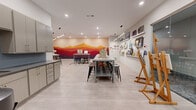 Located at 350 Old Hickory Blvd in Nashville, TN, this single-floor art studio offers a well-appointed, open-plan creative workspace designed to accommodate group classes and individual artistic sessions alike. The studio's most striking feature is a large, vivid mural spanning an entire accent wall, depicting layered mountain silhouettes in a warm gradient of deep burgundy, red, orange, and gold — an inspiring backdrop that sets the tone for the entire space.

The main studio floor features light-toned tile flooring and is illuminated by a combination of recessed ceiling lights and linear pendant fixtures, ensuring consistent, bright lighting across all work areas. Several sturdy worktables paired with black bar-height stools are arranged throughout the room, each station equipped with wooden tabletop easels to accommodate painters and artists of varying skill levels. Larger freestanding wooden easels are also positioned throughout the space for more expansive works.

Along one wall, tiered display shelves showcase an array of completed artwork, serving both as inspiration and a gallery of past student creations. On the opposite side, gray cabinetry provides organized storage for art supplies and materials. A dedicated storage area with lockers offers additional supply management. Decorative area rugs anchor key workstations, adding warmth and defining the layout within the open floor plan.