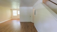 This two-floor space presents a straightforward, functional design with clean white walls and warm wood-look flooring that runs throughout the main level, creating a cohesive and inviting atmosphere. The main living area is open and well-proportioned, offering a sense of spaciousness that lends itself to flexible furniture arrangements. Natural light filters in through a front-facing window adjacent to the entry door, brightening the space and lending an airy quality to the interior. The textured ceiling adds a subtle architectural detail, while the smooth white walls provide a neutral backdrop throughout. A utility panel is visible along one wall, and interior doors lead to additional areas of the home, suggesting a practical and organized floor plan. The overall ambiance is clean and neutral, making the space highly adaptable for a variety of uses. With two floors, there is ample room for distinct living and private zones, providing both functionality and versatility for its occupants.