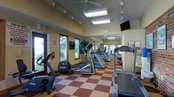 Located at 300 Valley Heights Drive in Williamsport, Pennsylvania, this fitness center offers a thoughtfully designed single-floor exercise facility built to support an active lifestyle. The gym features a generous open layout with a distinctive checkered tile floor in warm red and tan tones, lending the space a clean and energetic atmosphere. A row of cardio equipment — including treadmills, ellipticals, and stationary bikes — is arranged along the length of the room, with additional strength and cardio machines visible toward the rear. Large windows line the walls, flooding the interior with natural light and providing pleasant views of the surrounding wooded landscape. The vaulted ceiling is equipped with ceiling fans, recessed skylights, and track lighting, ensuring the space remains bright and well-ventilated throughout. A wall-mounted flat-screen television is positioned for easy viewing during workouts. Full-length mirrors along one wall visually expand the space and aid in exercise form. Safety features such as a fire extinguisher and clearly marked exit signage are prominently displayed. A bulletin board near the entrance provides a place for notices and community information. The overall ambiance is welcoming, functional, and well-maintained, making it a practical and comfortable space for daily fitness routines.