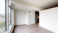 This well-appointed single-floor unit showcases a clean, modern design throughout, with light wood-tone flooring that flows seamlessly across the living, bedroom, and kitchen areas, creating a cohesive and contemporary aesthetic.

The open-concept living area is bright and airy, with neutral white walls that provide a versatile backdrop. It connects directly to the kitchen, which is outfitted with dark charcoal cabinetry, light countertops, and a full suite of stainless steel appliances including a refrigerator, electric range, built-in microwave, and dishwasher. Upper and lower cabinet storage is generously provided, along with an undermount sink and modern faucet.

The bedroom is a standout feature of the unit, boasting expansive floor-to-ceiling windows that frame sweeping panoramic views of the surrounding cityscape and greenery, flooding the room with abundant natural light. The space is well-proportioned and versatile.

The bathroom is finished to a high standard, featuring large-format tile flooring, dark tile surround in the soaking tub and shower combination, a sleek floating double vanity with integrated sink, backlit mirror, and brushed nickel hardware, all contributing to a refined, spa-like atmosphere.

Overall, the unit presents a polished and functional living environment with modern finishes and thoughtful design details throughout.