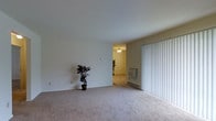 This single-floor space presents a clean, functional layout with a neutral color palette throughout. The main living area features wall-to-wall carpet in a warm beige tone, smooth white ceilings, and plain walls that provide a blank canvas for personalization. A doorway connects to an adjacent room, which is equipped with a ceiling fan and light fixture, adding practical overhead lighting and air circulation. Along one wall, a baseboard heating unit ensures efficient climate control. Vertical blinds cover what appears to be a sliding glass door, allowing for adjustable natural light and potential outdoor access. The overall ambiance is understated and practical, with minimal architectural detailing but a spacious, open feel. The consistent flooring and wall finishes throughout create a cohesive aesthetic. A decorative potted plant adds a touch of greenery to the otherwise neutral interior. The space is well-suited for straightforward furnishing and offers a functional foundation for comfortable everyday living.