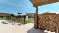 This outdoor community space is thoughtfully designed to encourage social gathering and recreational activity. At its centerpiece stands a sturdy wooden pergola with a warm reddish-brown finish, providing partial shade over a built-in outdoor kitchen equipped with two stainless steel grills set into a stone-faced counter. A picnic table positioned beneath the pergola offers a convenient dining area for residents enjoying outdoor meals. The pergola's open lattice roof allows natural light to filter through while framing views of the surrounding grounds and brick residential buildings.

Adjacent to the grilling area, a concrete patio extends toward a black wrought iron-fenced courtyard with well-maintained green lawn. Within this fenced area, lightweight seating chairs are arranged near the entrance, providing a relaxed spot to enjoy the outdoors. A posted information board near the gate outlines community rules for the space.

A playground area is visible in the background, complete with a green shade umbrella, making it a functional space for families. The grounds are maintained with neatly trimmed grass, and the open-sky setting ensures ample natural light throughout the day, contributing to an inviting and active outdoor environment.