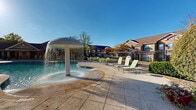 Located at 2000 Central Parkway in Montgomery, Alabama, this outdoor amenity area presents a well-appointed resort-style pool complex designed for relaxation and recreation. The centerpiece is a generously sized freeform swimming pool with crystal-clear blue water, accessible via stainless steel entry rails and surrounded by a broad, stamped concrete deck that provides ample space for lounging and socializing. A standout architectural feature is the mushroom-style water fountain, which cascades water in a dramatic curtain effect, adding both visual interest and a cooling atmosphere to the space. Complementing the main pool, a smaller wading or spa pool is also present nearby. The deck is outfitted with multiple green sling-style chaise lounges and side tables, thoughtfully arranged throughout the patio. A charming wooden pergola anchors one end of the sunning area, flanked by tall palm trees and lush landscaping that lend a tropical, resort-like character. A craftsman-style pool house with stone accents, a pitched roof, and covered portico serves as a functional support structure, offering restroom facilities and additional shade. The entire area is enclosed by ornamental iron fencing, ensuring privacy and security. Under open skies, the space enjoys abundant natural light, creating a bright and inviting outdoor retreat.