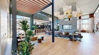 Located at 4692 E Northfield Blvd in Denver's Central Park community, this expansive single-floor lounge spans a generously sized footprint designed to accommodate a variety of social and collaborative functions. The interior architecture is defined by soaring double-height ceilings, warm light wood flooring, exposed structural steel framing, and a mix of wood-slat ceiling accents and exposed brick walls that lend an upscale yet approachable aesthetic throughout.

The main lounge area centers around a dramatic floor-to-ceiling stacked stone fireplace flanked by sculptural pendant lighting with gold and white globe fixtures. Multiple seating arrangements—featuring leather armchairs, plush sofas, and wooden accent tables—are distributed throughout, creating distinct social zones. Expansive floor-to-ceiling windows and clerestory glazing flood the space with natural light and provide outdoor views of the surrounding community and pool area.

A dedicated dining and lounge nook features geometric patterned tile flooring, a long communal wood table with leather chairs, amber glass globe pendants, and a wet bar with dark tile backsplash. Adjacent to this, a glass-walled conference or work room offers a quieter setting. A window-facing counter workspace with bar seating overlooks the pool, rounding out the functional areas of this well-appointed lounge.
