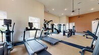 This fitness center offers a generously sized, single-floor workout facility designed to accommodate a full range of exercise activities. The main workout area features soaring ceilings with recessed lighting and pendant ceiling fans, creating an open and airy atmosphere. Large windows line the walls, flooding the space with natural light and offering views of the surrounding outdoors. The flooring consists of a distinctive striped carpet pattern in contrasting tan and dark blue tones, defining different workout zones throughout the room. Cardio equipment, including multiple treadmills and an elliptical, is positioned near the windows, while a comprehensive selection of strength training machines occupies the central floor. A dedicated free weights area features a full rack of dumbbells, an adjustable bench, and a Smith machine, all complemented by full-length mirrors and a wall-mounted flat-screen television. An accent wall in a warm terracotta tone adds a bold design element, anchoring the free weights zone. An adjoining room, accessible through a doorway, provides additional space for stretching, group exercise, or alternative training. The overall ambiance is clean, modern, and purposefully designed to support a well-rounded fitness experience.