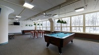 This single-floor recreational space is designed for leisure and social activity, centered around multiple full-sized pool tables illuminated by classic green pendant lights suspended from above. The room offers an open, airy layout with neutral carpet flooring that extends throughout the space, providing a comfortable and relaxed atmosphere.

Large multi-pane windows line the walls, flooding the interior with natural light and offering sweeping views of a wooded landscape, particularly striking in winter when snow dusts the surrounding trees. The exposed ceiling features a combination of recessed fluorescent lighting and ceiling fans, contributing to the industrial-casual aesthetic of the room.

A casual dining area with bar-height tables and metal stools is positioned near the windows, offering a comfortable spot to relax between games. The white walls and bright lighting keep the space feeling open and inviting. Additional game tables are visible toward the back of the room, suggesting the space is well-equipped for a variety of recreational activities. Overall, the room strikes a balance between functional design and a welcoming, social ambiance ideal for group gatherings and recreational use.