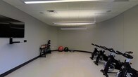 This single-floor fitness space is designed to support a well-rounded workout experience in a clean, functional environment. The room features a dropped tile ceiling equipped with bright, evenly distributed fluorescent lighting that ensures excellent visibility throughout the space. The walls are finished in a neutral gray tone, complemented by dark baseboards and a smooth, light-colored floor that is easy to maintain and suitable for a variety of exercise activities. The layout is open and spacious, allowing for comfortable movement between stations. A rack of kettlebells of varying weights is positioned along one wall, providing a versatile free-weight option for strength and conditioning training. Nearby, an assortment of stability and medicine balls, including a large red exercise ball, a black medicine ball, and a BOSU balance trainer, offer options for core training and functional fitness work. On the opposite side of the room, a row of stationary cycling bikes provides a dedicated cardio zone. The overall ambiance is practical and no-frills, prioritizing usability and a distraction-free environment ideal for focused training sessions.