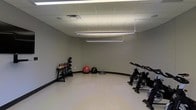 This single-floor fitness space is designed to support a well-rounded workout experience in a clean, functional environment. The room features a dropped tile ceiling equipped with bright, evenly distributed fluorescent lighting that ensures excellent visibility throughout the space. The walls are finished in a neutral gray tone, complemented by dark baseboards and a smooth, light-colored floor that is easy to maintain and suitable for a variety of exercise activities. The layout is open and spacious, allowing for comfortable movement between stations. A rack of kettlebells of varying weights is positioned along one wall, providing a versatile free-weight option for strength and conditioning training. Nearby, an assortment of stability and medicine balls, including a large red exercise ball, a black medicine ball, and a BOSU balance trainer, offer options for core training and functional fitness work. On the opposite side of the room, a row of stationary cycling bikes provides a dedicated cardio zone. The overall ambiance is practical and no-frills, prioritizing usability and a distraction-free environment ideal for focused training sessions.