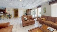 This well-appointed single-floor communal space is designed for comfort and socializing, offering a seamless flow between its distinct functional areas. The interior is unified by striking coffered tile ceilings with recessed lighting, warm yellow wainscoted walls, and large-format light tile flooring that carries throughout the entire space.

The expansive living area is anchored by plush leather seating, an ornate carved wood coffee table, and a large flat-screen television. Multiple arched windows and glass doors flood the room with natural light while offering views of the surrounding outdoor area, including a pool and lush palm trees.

A dedicated dining area accommodates a full-sized wooden table with seating for several guests, complemented by decorative indoor trees and a bookshelf, creating a warm and inviting atmosphere. Large windows frame views of the outdoor pool deck, enhancing the connection between indoor and outdoor spaces.

The kitchen is fully equipped with rich honey-toned cabinetry, dark granite countertops, stainless steel appliances including a refrigerator and range, and an ornamental coffered ceiling detail. A secondary kitchenette area near the pool-facing exit provides additional counter space and refrigeration, ideal for gathering events. Direct glass door access to the pool area makes this space a true hub for community activity.