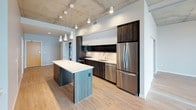 This contemporary urban unit showcases an industrial-modern aesthetic defined by striking exposed concrete ceilings that extend throughout the open living areas, creating an elevated, loft-like ambiance. Light hardwood flooring flows seamlessly across the main living spaces, complementing the clean, neutral color palette.

The kitchen is a standout feature, equipped with a full suite of stainless steel appliances, including a French door refrigerator, range, microwave, and dishwasher. Dark wood cabinetry is paired with crisp white upper cabinets and light countertops, while a central island with a white quartz surface provides additional workspace and storage. Pendant lighting adds warmth above the island.

The open living area benefits from expansive floor-to-ceiling windows that frame broad city views, flooding the space with natural light. Built-in shelving offers practical storage and display options. Bedrooms continue the floor-to-ceiling window theme, with one featuring plush carpeting and sweeping urban vistas framed by exposed concrete columns.

Two bathrooms serve the unit — one featuring a soaking tub with tile surround and a dark wood vanity with a white countertop, and a second, more compact bathroom with a matching vanity and grab bar. Both bathrooms feature hardwood-style flooring and clean, minimalist finishes throughout.