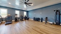 Located at 4420 Taylor Rd in Nashville, Tennessee, this well-appointed fitness center offers a comprehensive workout environment within a single, open-plan exercise room. The space is characterized by its elevated tray ceiling finished with white shiplap paneling, a large ceiling fan for air circulation, and recessed lighting that keeps the room bright and inviting. Bold blue-gray walls provide a clean, motivating backdrop throughout.

Natural light flows in through multiple white-trimmed windows positioned along the walls, offering views of the surrounding area while maintaining an open, airy atmosphere. The floor is covered in warm-toned, wood-style vinyl plank flooring that is both durable and easy to maintain.

Equipment is thoughtfully arranged to maximize usable floor space. The cardio zone features treadmills, an elliptical, and a stationary bike, while the strength training area includes a full power rack with a barbell and weight plates, a multi-use cable machine, and an adjustable workout bench. A well-stocked three-tier dumbbell rack provides a full range of free weights. Functional training accessories such as kettlebells, medicine balls, stability balls, and resistance tools are organized along a mirrored wall, supporting a wide variety of workout styles. The open central floor area allows ample room for stretching or bodyweight exercises.
