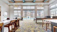 This single-floor clubhouse is a thoughtfully designed amenity space that combines social, recreational, and functional areas into one cohesive environment. The interior is anchored by soaring double-height ceilings with teal-accented coffered beams, and dramatic sputnik-style chandeliers that serve as bold focal points throughout the space.

A generously sized game and lounge area dominates one wing, featuring a billiards table, high-top seating, and a vibrant patterned carpet in grey and yellow tones that flows continuously throughout. Floor-to-ceiling glass walls and transom windows flood the space with natural light and offer unobstructed views of the outdoor pool and lounge deck beyond.

A communal kitchen and dining area provides a large white-topped island with bar-height wooden seating, open shelving, and appliances suited for casual gatherings. A large-scale bison mural and decorative sculptural accents lend the space a distinctive, nature-inspired character.

The leasing and reception area features a sleek, upholstered front desk with mirrored upper panels and an adjacent lounge sofa for a welcoming first impression. Nearby, intimate private meeting pods lined in deep blue upholstery offer semi-enclosed seating for focused conversations or individual work, complementing the overall flexible, community-oriented design.