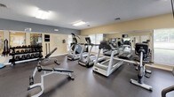 This fitness center offers a thorough and well-organized workout environment designed to accommodate a wide range of exercise routines. The space is laid out on a single open floor, maximizing usable area and allowing for easy navigation between equipment zones.

The cardio section is anchored by a row of Matrix treadmills and elliptical trainers, including a True-brand elliptical, all positioned to face the room's large windows that bring in ample natural light and offer views of the surrounding greenery. A wall-mounted flat-screen television provides entertainment during workouts.

A full-length mirror spans one wall, visually expanding the space and allowing users to monitor their form throughout their routines. The free weight area features a comprehensive dumbbell rack with a wide range of weights, a multifunctional adjustable bench, and an assortment of exercise balls and resistance bands for varied training. A cable-based multi-station strength machine rounds out the strength training offerings.

The flooring is covered in durable dark rubber, providing safety and support underfoot. Overhead fluorescent lighting ensures the space remains well-lit throughout. The walls are painted in neutral warm tones with gray accents, contributing to a clean and focused atmosphere. A textured ceiling and multiple windows complete the practical, no-frills design of this functional fitness facility.