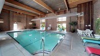 This indoor aquatic facility is a well-appointed space defined by its distinctive architectural character and functional design. The centerpiece is a generously sized lap pool with lane dividers, its clear turquoise water reflecting the warm tones of the surrounding environment. The pool is enclosed within soaring brick walls that lend a sturdy, classic aesthetic to the space, while exposed wooden beams and a metal-clad ceiling overhead add an industrial-meets-rustic charm. Tall windows positioned along the upper walls allow natural light to filter in, brightening the space and creating an inviting atmosphere throughout the day. Overhead, large cylindrical HVAC ducts run the length of the facility, ensuring climate control year-round. The broad concrete pool deck provides ample space for movement and relaxation, and is accented by lush potted tropical plants and palms strategically placed around the perimeter, softening the industrial elements and adding a vibrant, resort-like feel. Seating areas with white chairs are available poolside. Access to the facility includes glass double doors from an adjacent area, with outdoor lounge chairs also available nearby. Recessed and pendant lighting supplements natural light for evening or low-light use, making this a fully functional, all-season aquatic space.