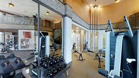 This fitness center combines functionality with thoughtful design, creating an inviting environment for a full range of workout activities. The space is distinguished by soaring ceilings that lend an open, airy atmosphere, complemented by warm, ambient pendant lighting that adds a modern industrial touch. Exposed brick detailing along the upper walls adds character and texture, while large glass panel partitions divide the space without sacrificing visual openness.

The strength training area is thoroughly outfitted with a wide selection of kettlebells arranged on tiered racks, cable machines, and a variety of resistance and functional training equipment. Mirrored walls enhance the sense of space and allow for proper form checks during exercise. The flooring transitions appropriately between zones, supporting both free weight and machine-based training.

Natural light filters through the space, and the warm-toned walls paired with polished finishes create an upscale yet practical atmosphere. The layout is well-organized, allowing for ease of movement between equipment stations. Whether focusing on strength, flexibility, or functional fitness, this facility provides a comprehensive and well-appointed space suited to a wide range of training needs.